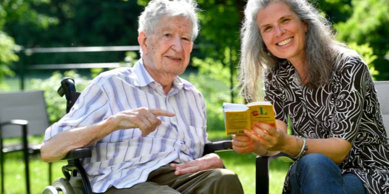 freiwilligenkoordinatorin und ein älter bewohner im rollstuhl sitzen im garten des muenchenstift