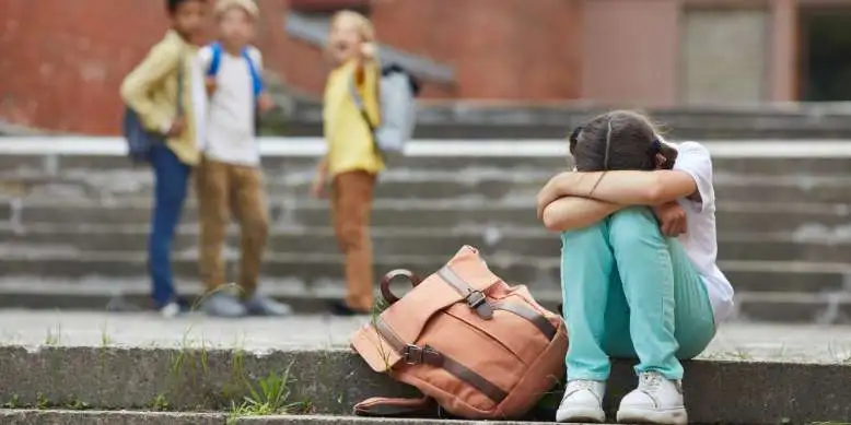 Mädchen sitzt auf einer Treppe, Kopf auf den Knien im Hintergrund steht eine Gruppe Kinder und spricht über das Mädchen